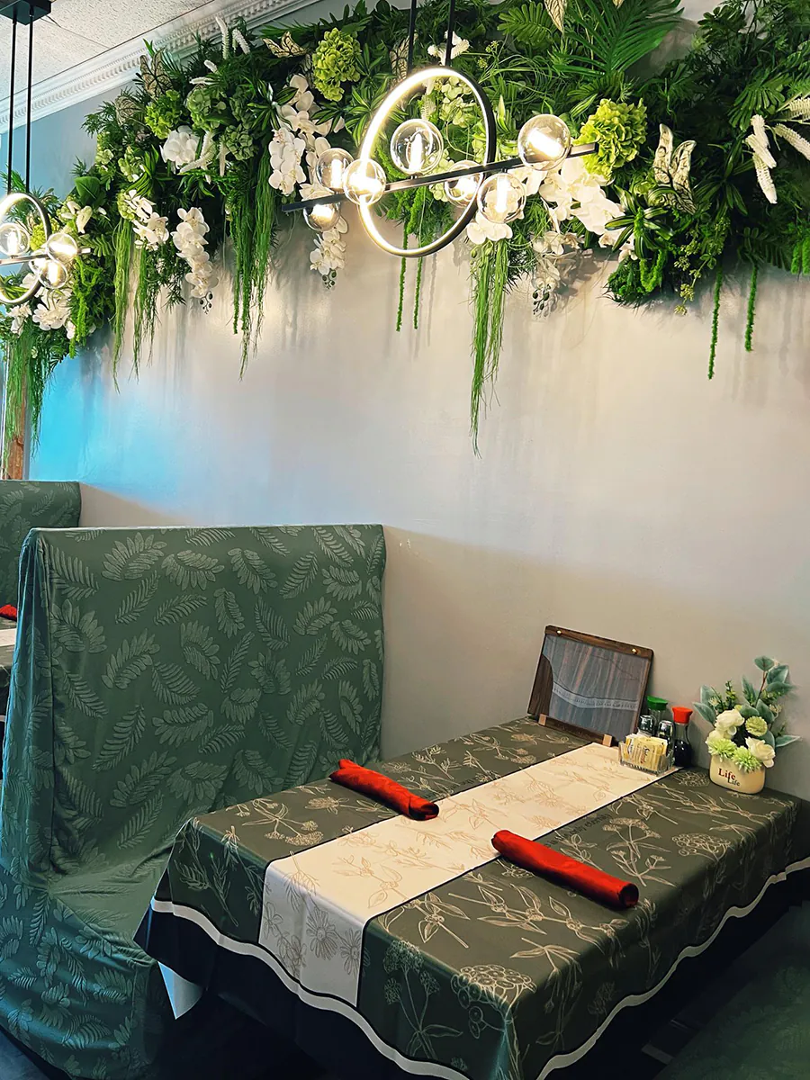 Restaurant interior with green leafy decor, hanging plants, and a dining table with a botanical-patterned tablecloth at J&J's Garden Kitchen，a Asian Restaurant in Fredericksburg